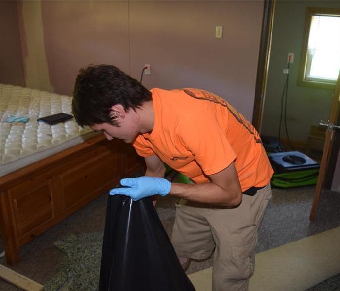 Young man in orange SERVPRO t-shirt with blue gloves removing wet damaged materials from bedroom